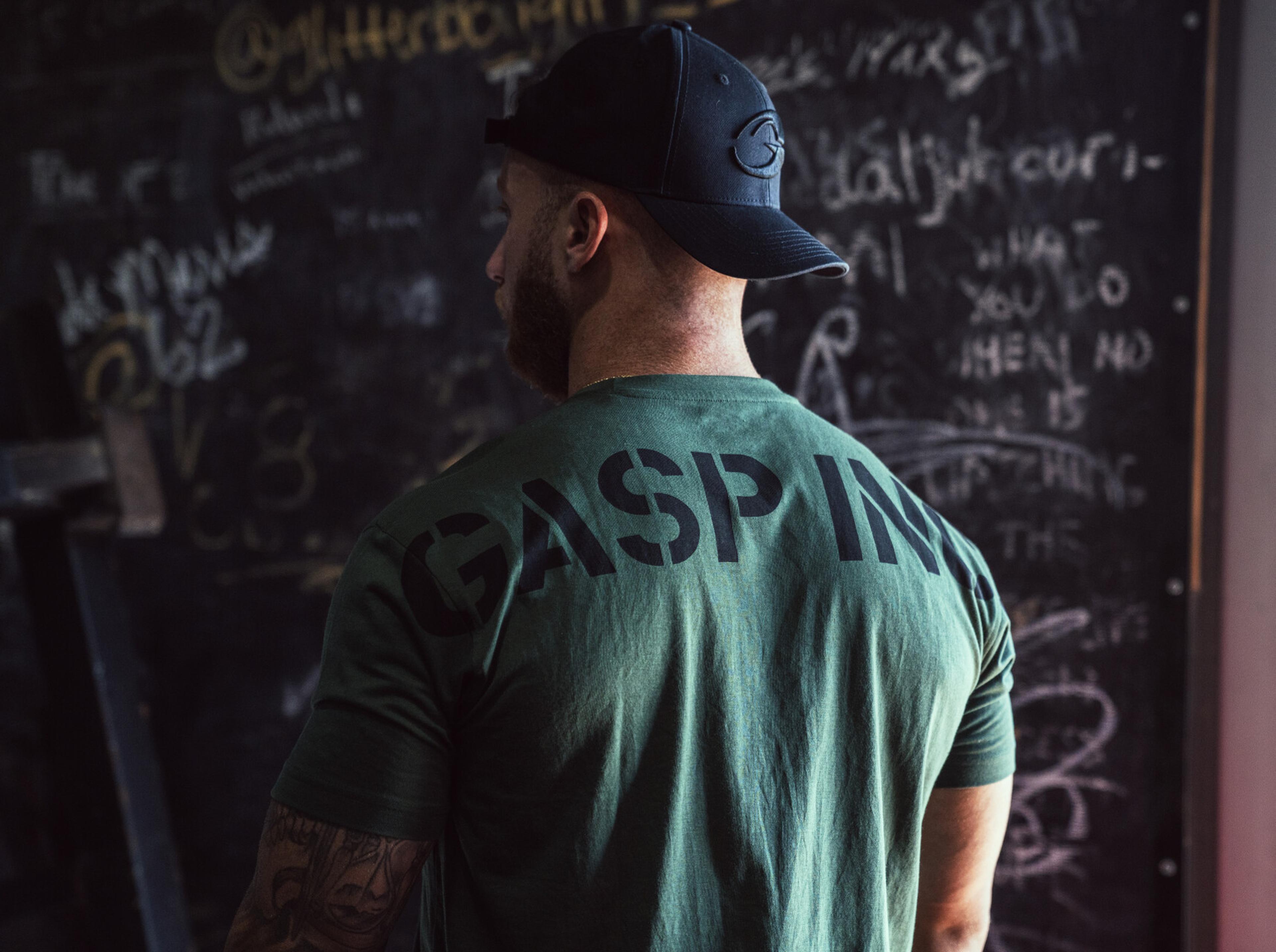 Man in a green "GASP INC" shirt and black cap stands in front of a chalkboard with various writings.
