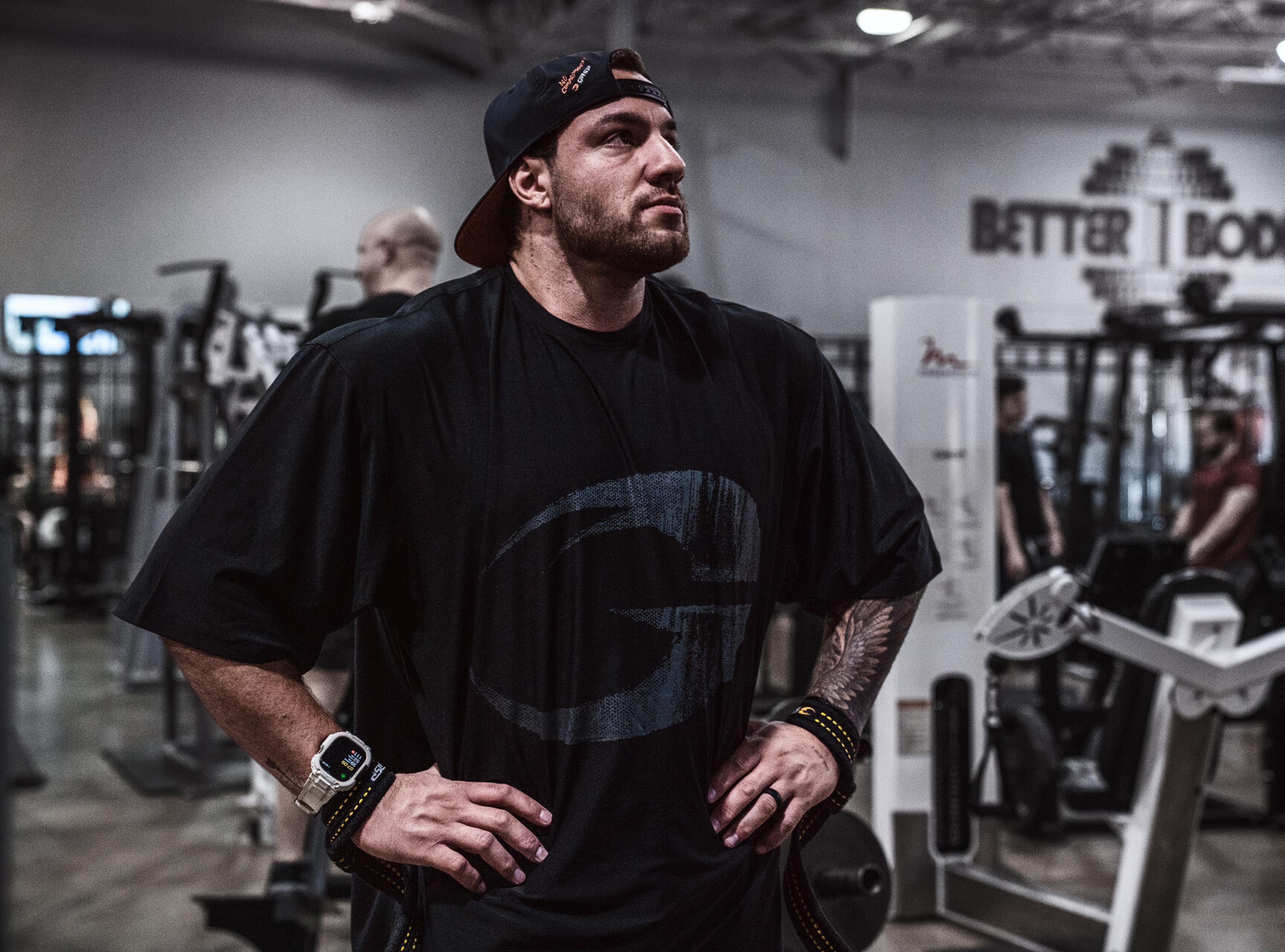 A man in a gym wearing a black T-shirt and cap, with hands on hips, stands confidently among exercise equipment.