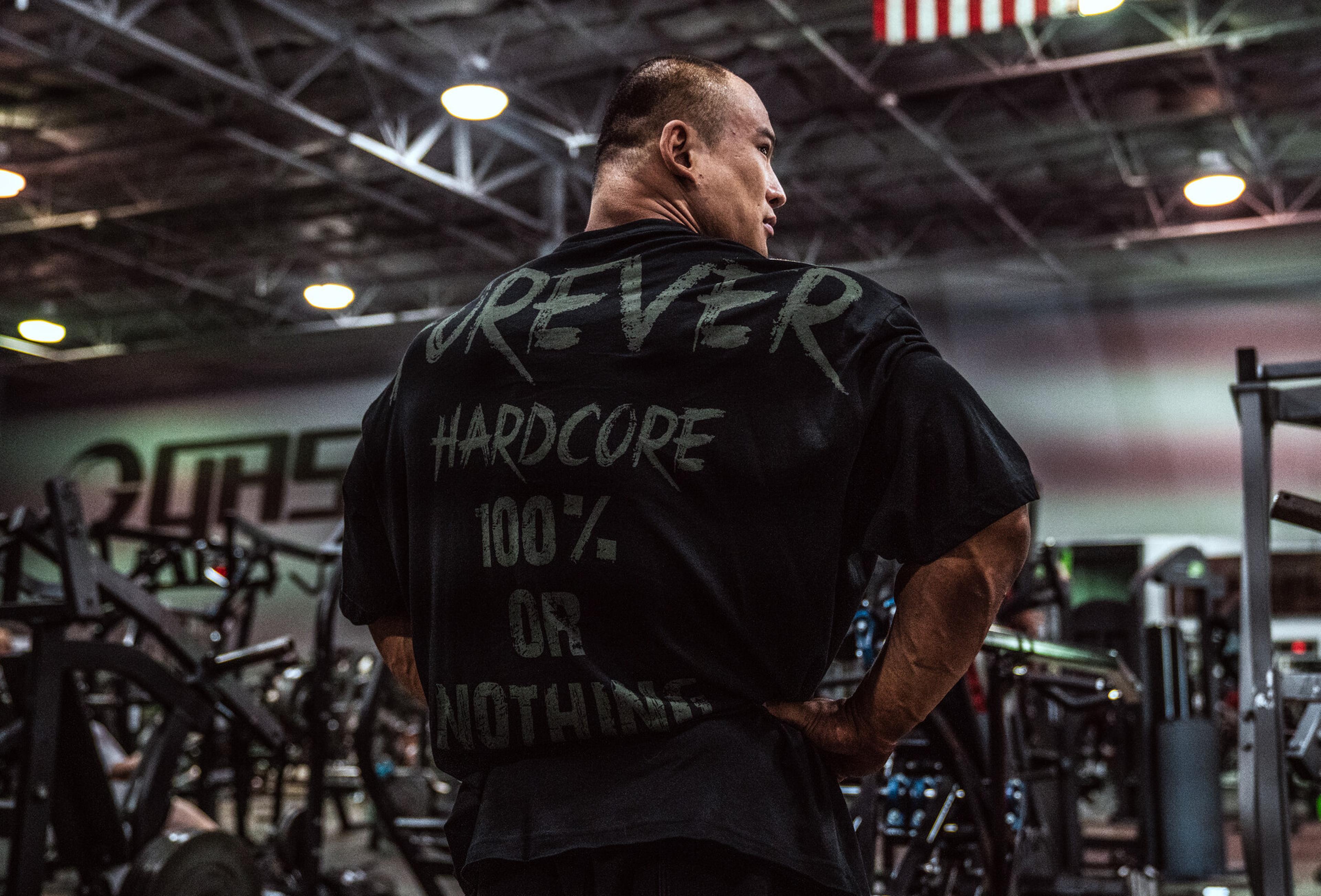 Man in a gym wearing a black shirt with "FOREVER HARDCORE 100% OR NOTHING" printed on the back, standing among workout equipment.