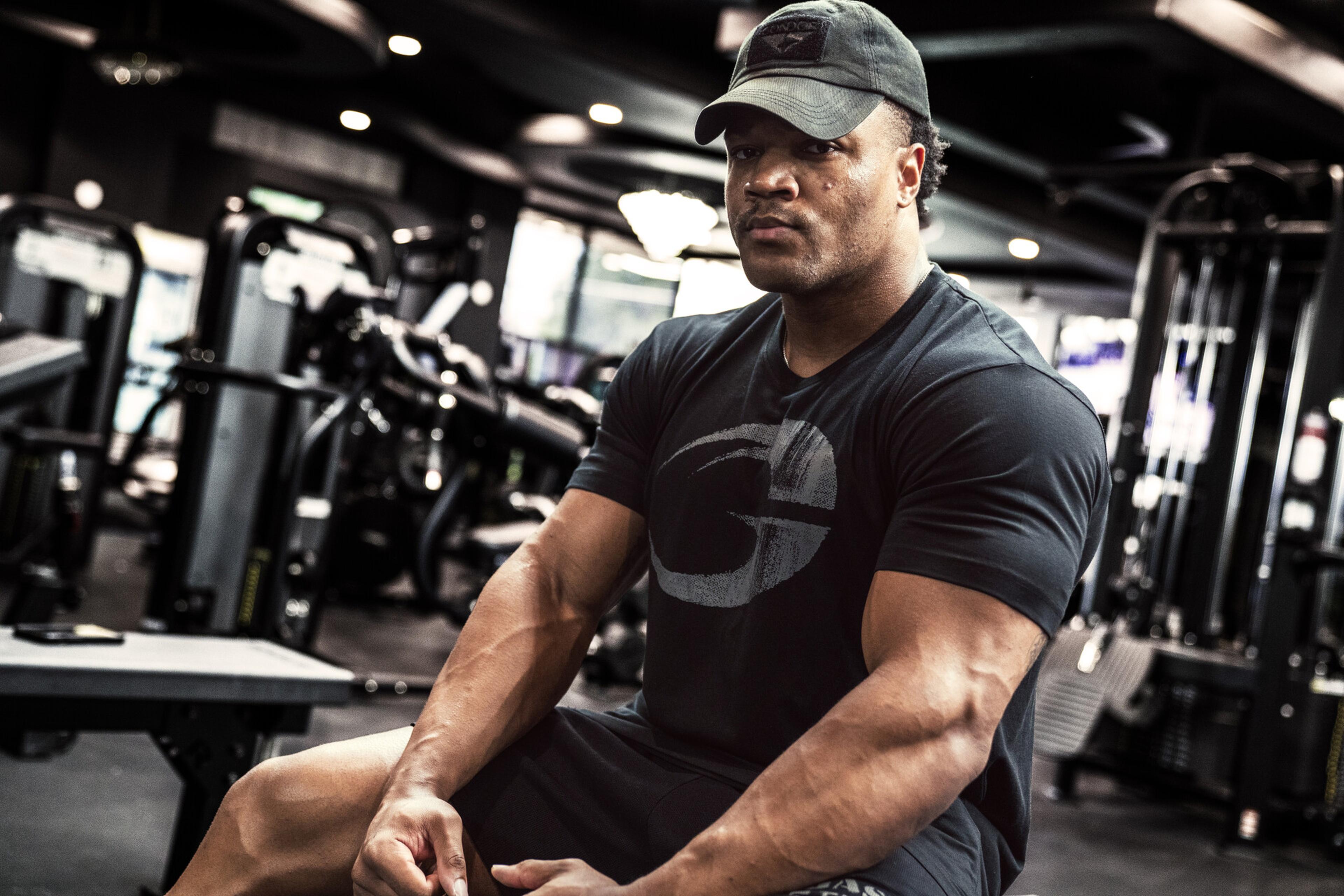 A muscular man in a black T-shirt and cap sits on a bench in a gym, surrounded by workout equipment.