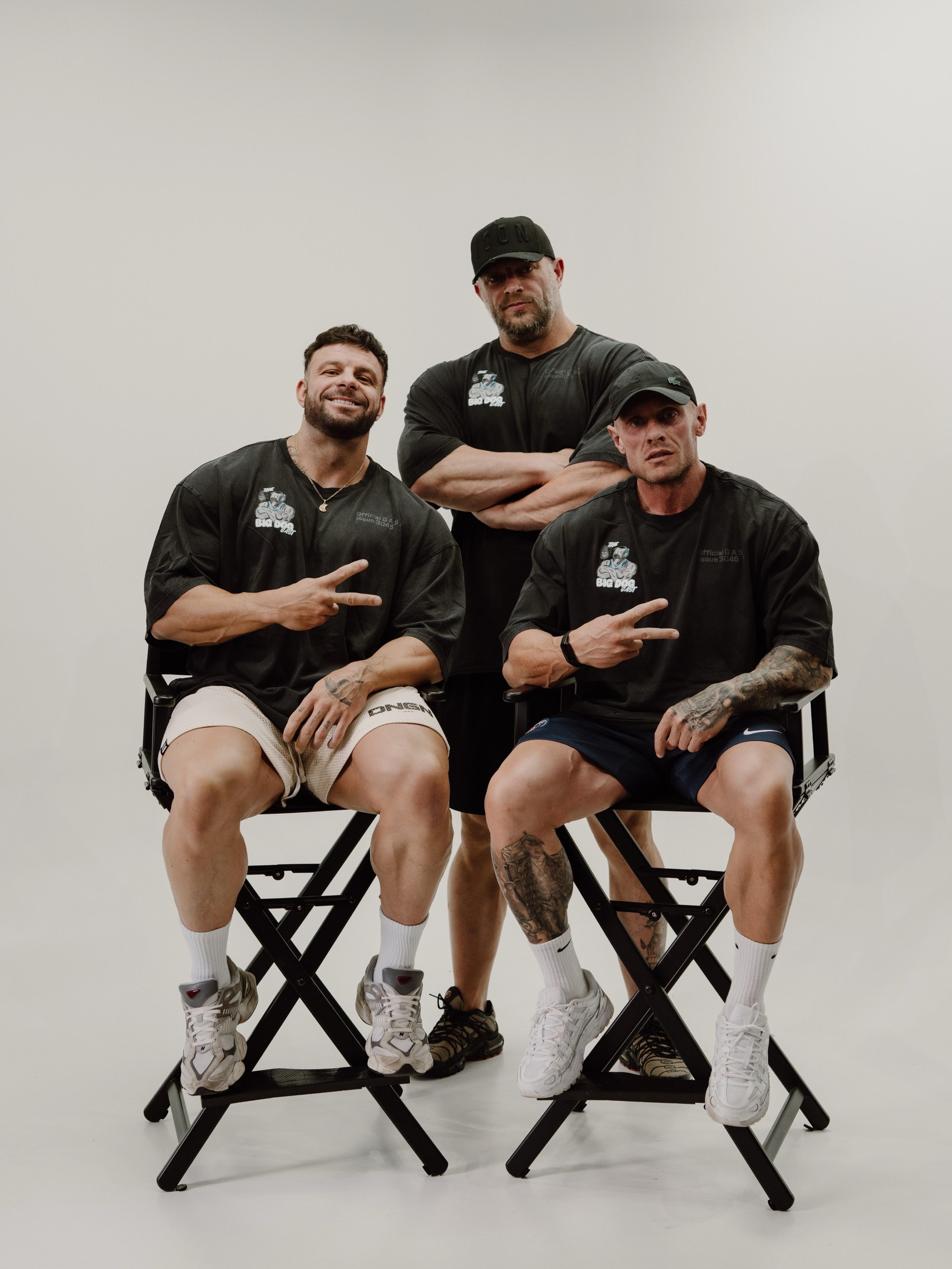 Three men in black shirts and shorts pose with peace signs on director chairs against a plain background.
