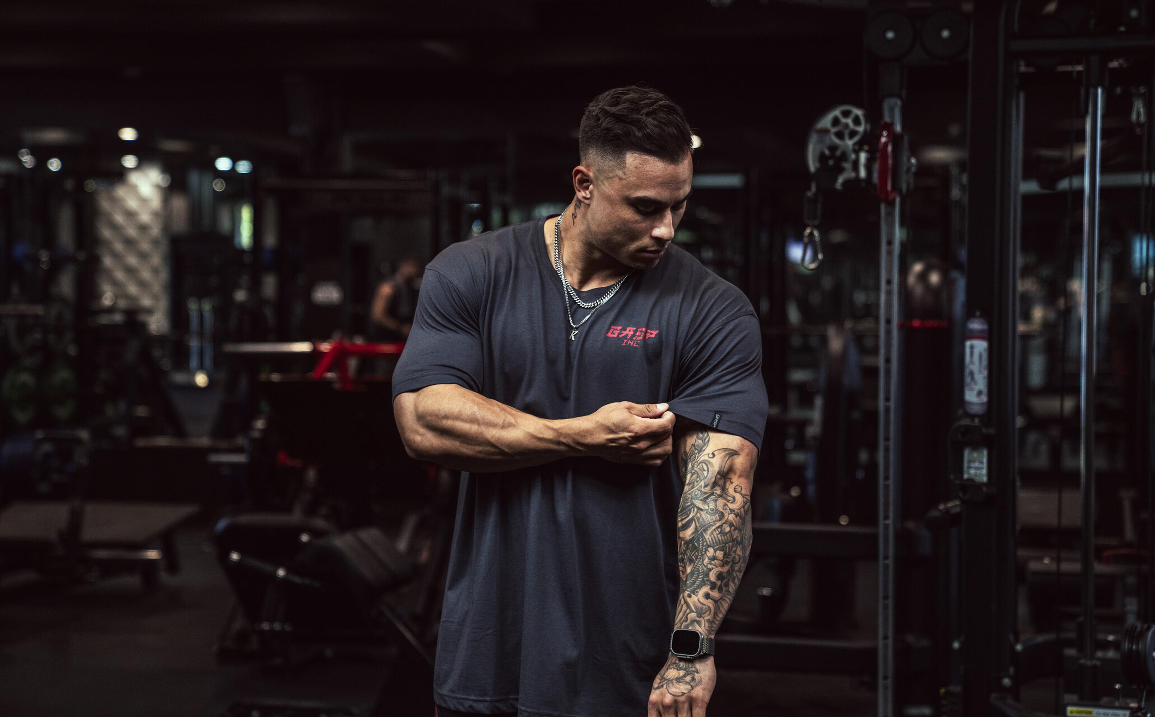 A man in a gym, wearing a dark shirt, looks at his tattooed arm. Gym equipment is visible in the background.