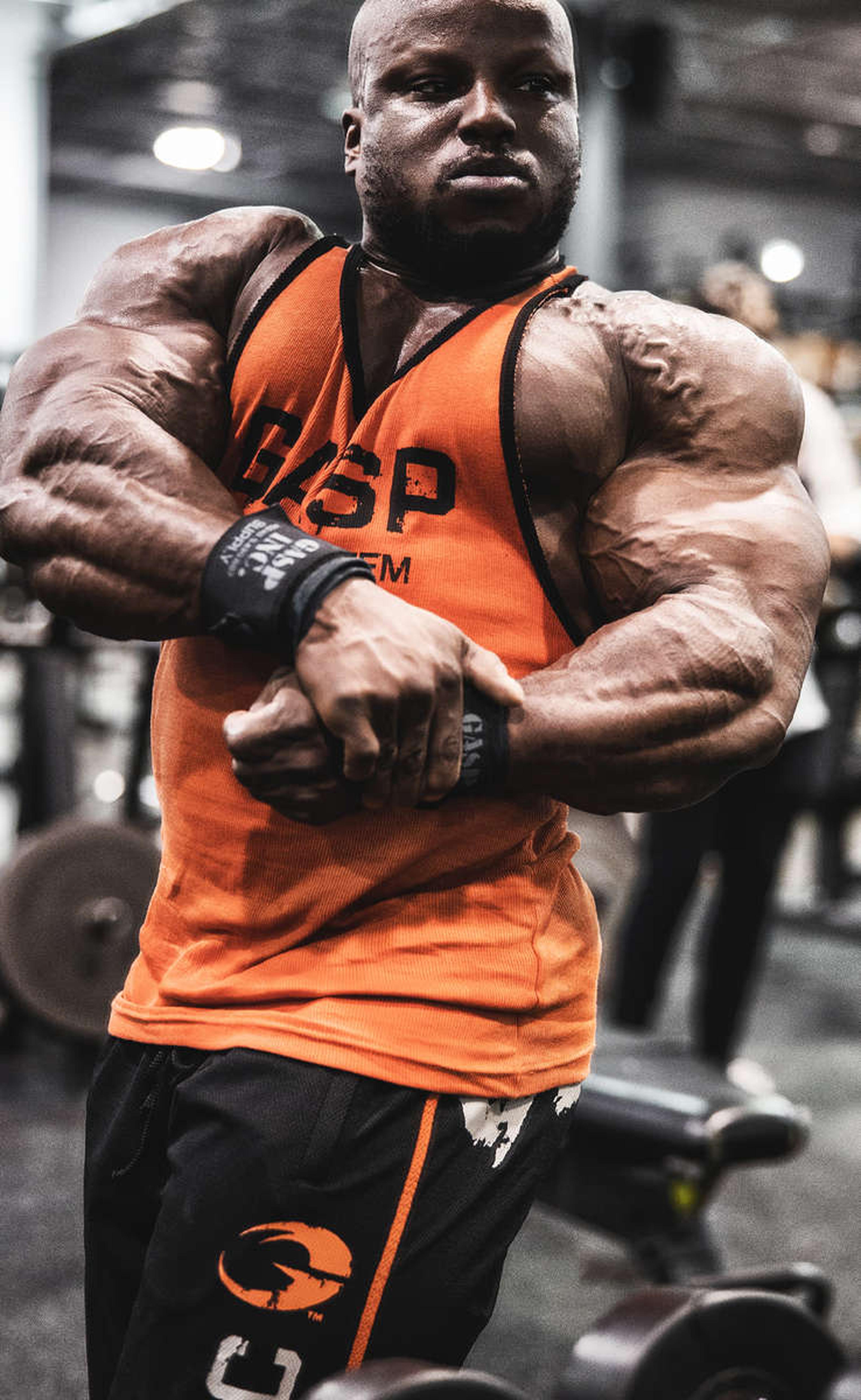 Bodybuilder in an orange tank top and black shorts flexing muscles in a gym, with weights in the background.