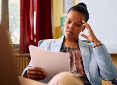 A woman in a blue blazer sits in an office, holding and reading a document, looking thoughtful.