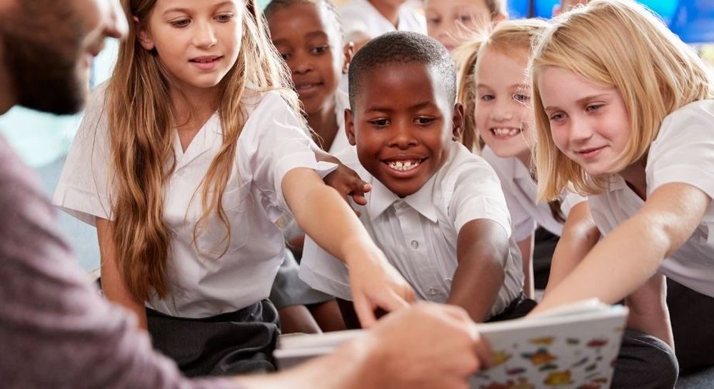 A group of smiling children in school uniforms eagerly reaching towards a book held by an adult.