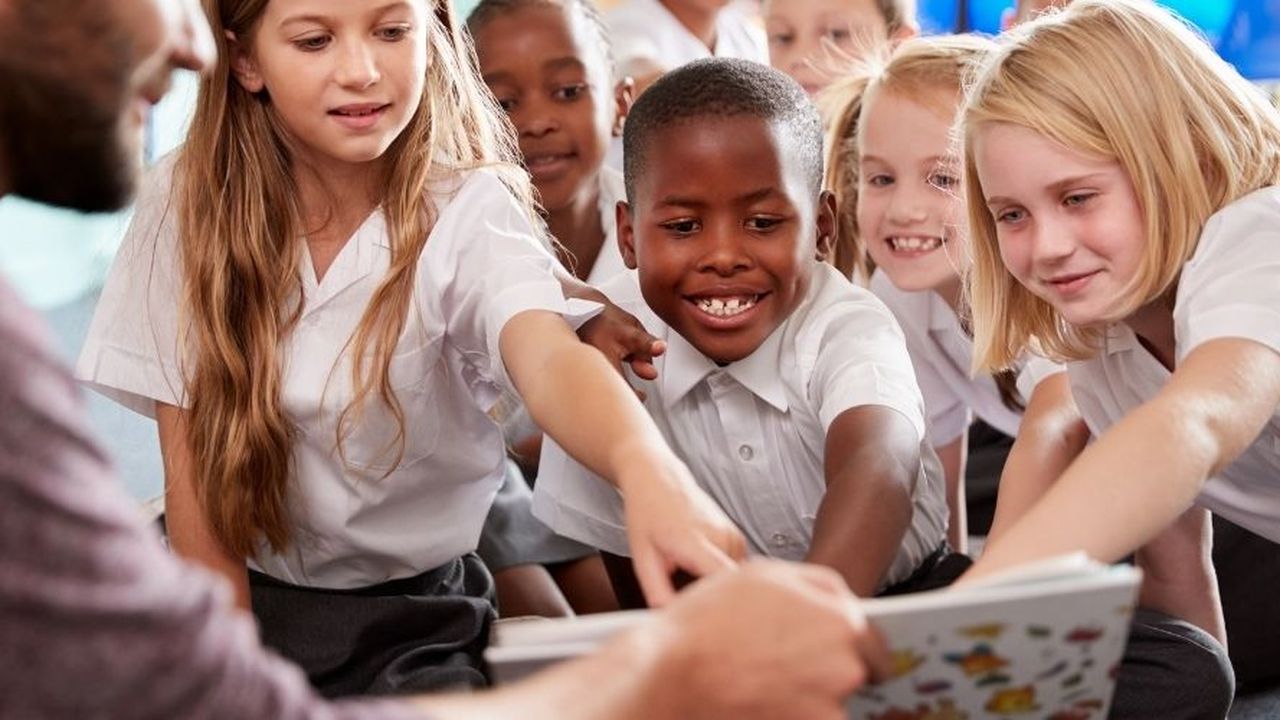 A group of smiling children in school uniforms eagerly reaching towards a book held by an adult.