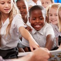 A group of smiling children in school uniforms eagerly reaching towards a book held by an adult.