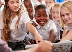A group of smiling children in school uniforms eagerly reaching towards a book held by an adult.