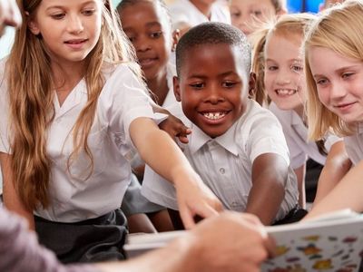 A group of smiling children in school uniforms eagerly reaching towards a book held by an adult.