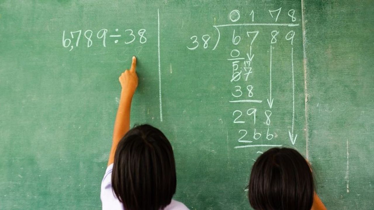 Two children solving a division problem on a green chalkboard, with one pointing at the equation "6,789 ÷ 38" written in chalk.