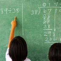 Two children solving a division problem on a green chalkboard, with one pointing at the equation "6,789 ÷ 38" written in chalk.