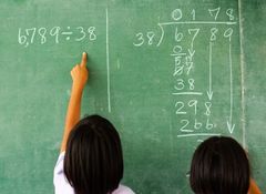 Two children solving a division problem on a green chalkboard, with one pointing at the equation "6,789 ÷ 38" written in chalk.