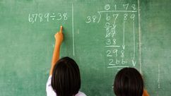 Two children solving a division problem on a green chalkboard, with one pointing at the equation "6,789 ÷ 38" written in chalk.