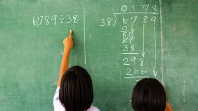 Two children solving a division problem on a green chalkboard, with one pointing at the equation "6,789 ÷ 38" written in chalk.