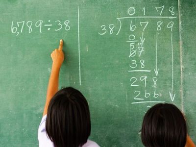 Two children solving a division problem on a green chalkboard, with one pointing at the equation "6,789 ÷ 38" written in chalk.