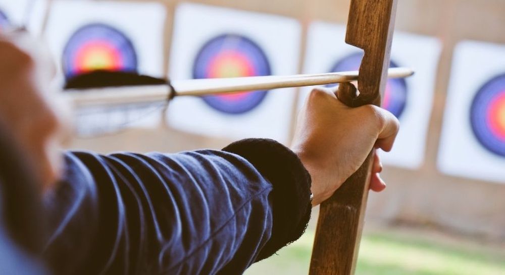 Close-up of a person drawing a bow, aiming at multiple archery targets in the background.