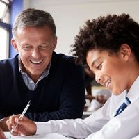 Teacher helping a smiling student with curly hair in a classroom, both looking at a notebook. Other students are visible in the background.