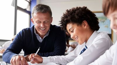 Teacher helping a smiling student with curly hair in a classroom, both looking at a notebook. Other students are visible in the background.