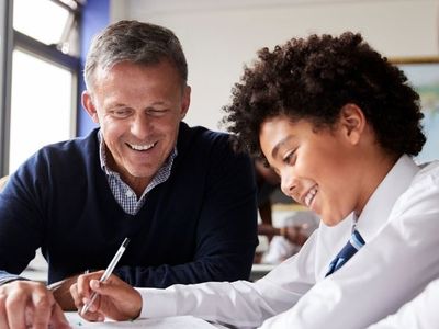 Teacher helping a smiling student with curly hair in a classroom, both looking at a notebook. Other students are visible in the background.