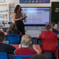 A teacher stands in front of a classroom with students seated at desks, using tablets. A screen displays colourful charts behind her.