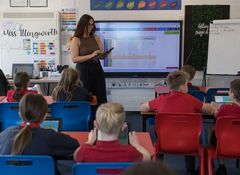 A teacher stands in front of a classroom with students seated at desks, using tablets. A screen displays colourful charts behind her.