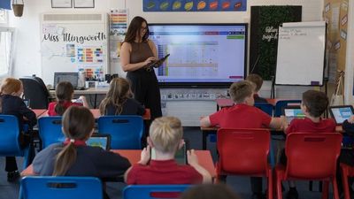 A teacher stands in front of a classroom with students seated at desks, using tablets. A screen displays colourful charts behind her.