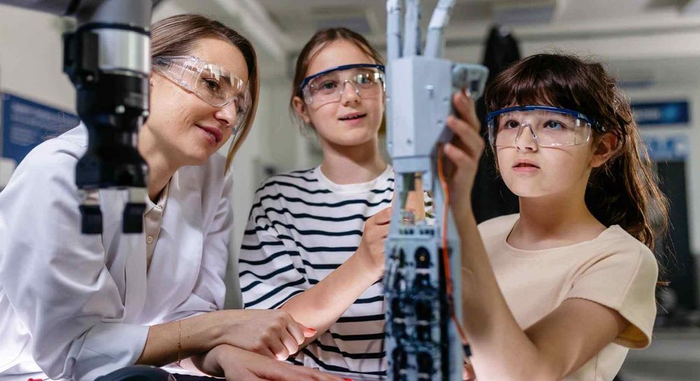 Two girls wearing safety goggles work on a robotic arm in a lab setting, guided by a woman in a white coat.