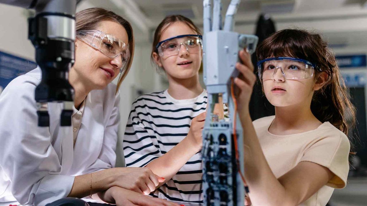 Two girls wearing safety goggles work on a robotic arm in a lab setting, guided by a woman in a white coat.
