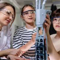 Two girls wearing safety goggles work on a robotic arm in a lab setting, guided by a woman in a white coat.