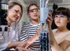 Two girls wearing safety goggles work on a robotic arm in a lab setting, guided by a woman in a white coat.