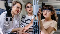 Two girls wearing safety goggles work on a robotic arm in a lab setting, guided by a woman in a white coat.