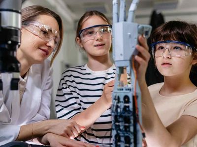 Two girls wearing safety goggles work on a robotic arm in a lab setting, guided by a woman in a white coat.