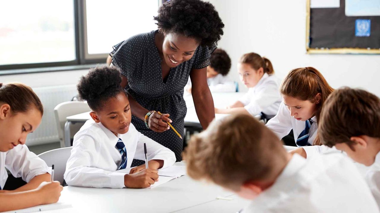 Teacher assisting students in a classroom. Students in uniforms focus on writing at their desks. Bright, well-lit room.
