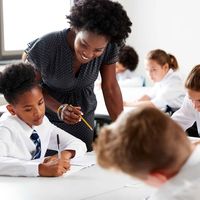 Teacher assisting students in a classroom. Students in uniforms focus on writing at their desks. Bright, well-lit room.