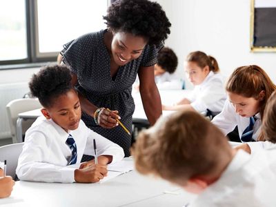 Teacher assisting students in a classroom. Students in uniforms focus on writing at their desks. Bright, well-lit room.