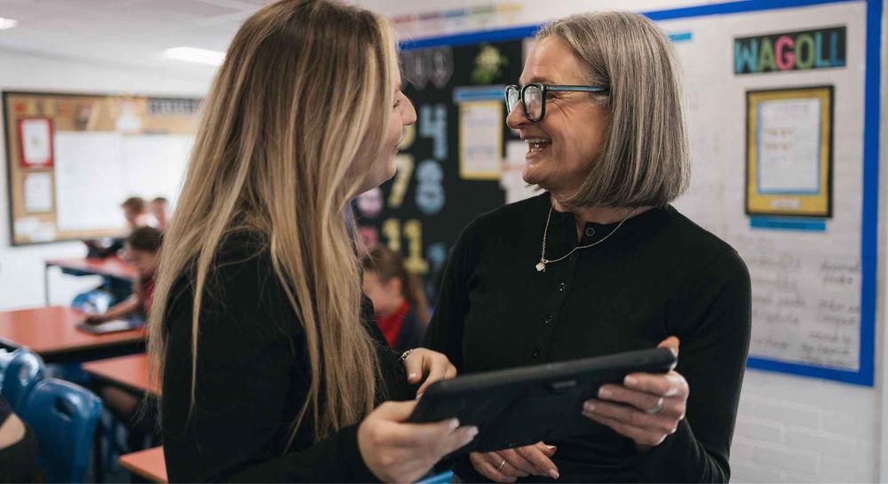 Two teachers chatting in a classroom holding an iPad.