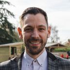 Smiling man with a neatly trimmed beard and short hair, wearing a checked blazer and white shirt, standing outdoors with a blurred background.