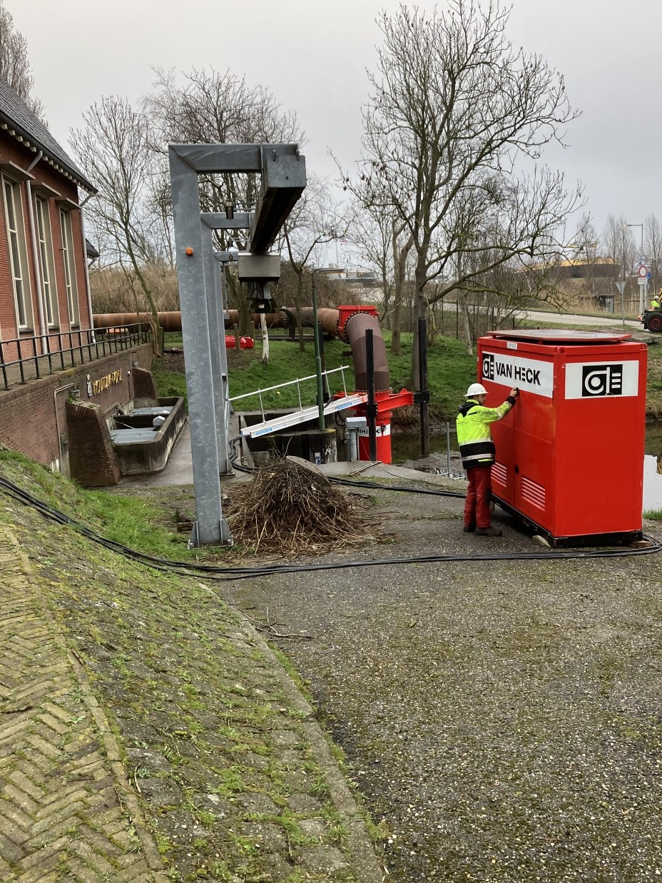 Opbouw van tijdelijke pompinstallatie bij een gemaal Monteur staat naast een rode container met opschrift "Van heck" in de nabijheid van een gemaal en roostergoedreiniger met een rode pomp op de achtergrond