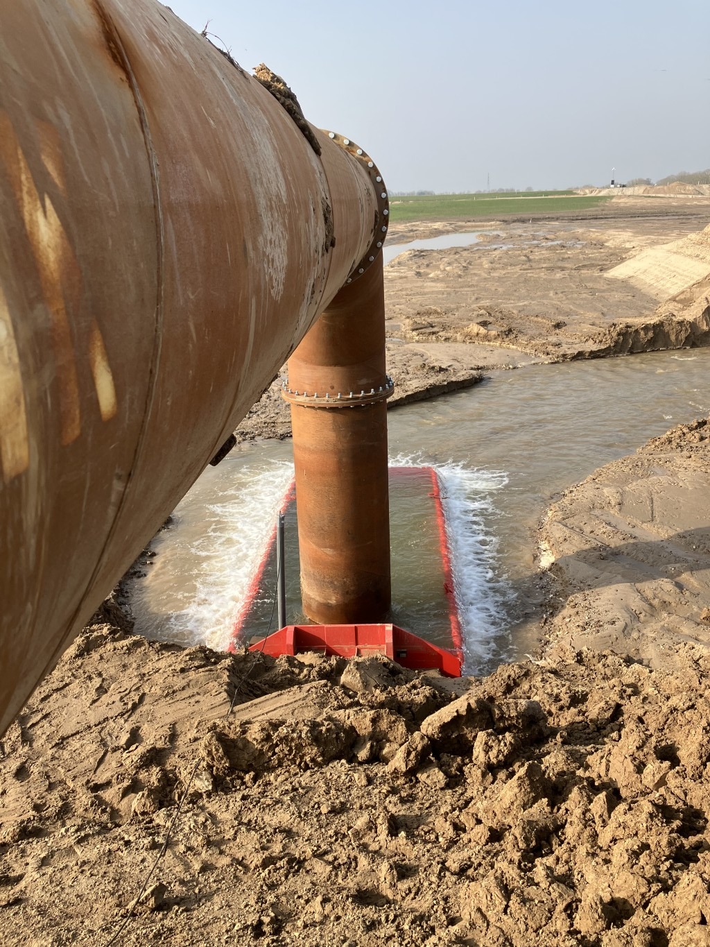 Uitstroomcontainer hevelinstallatie Stalen leiding die in een rode container staat waar water uitstroomt in een zanderige omgeving