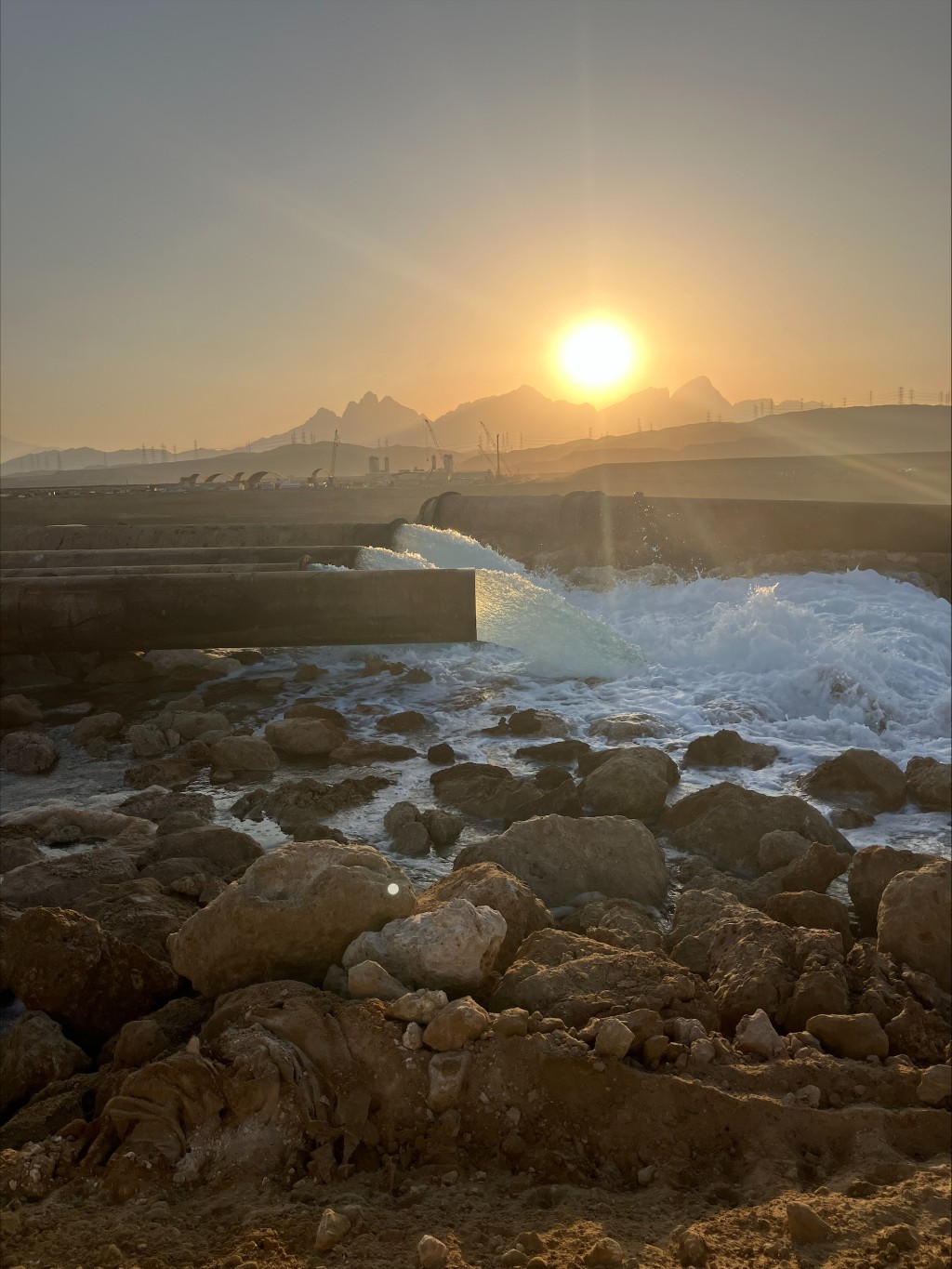 Leidingwerk in een woestijnomgeving waaruit een grote hoeveelheid water spuit onder grote kracht bij zonsondergang