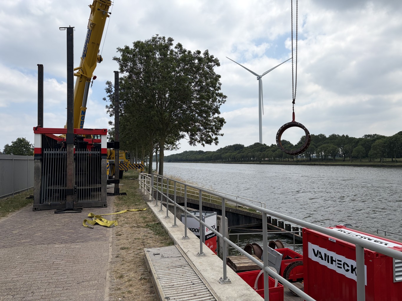 Kraag hijst een stuk leidingwerk naast een kade met daarop een zwarte unit die lijkt op een pomp. Daarnaast is een kanaal met een grijze lucht met daarop een windmolen