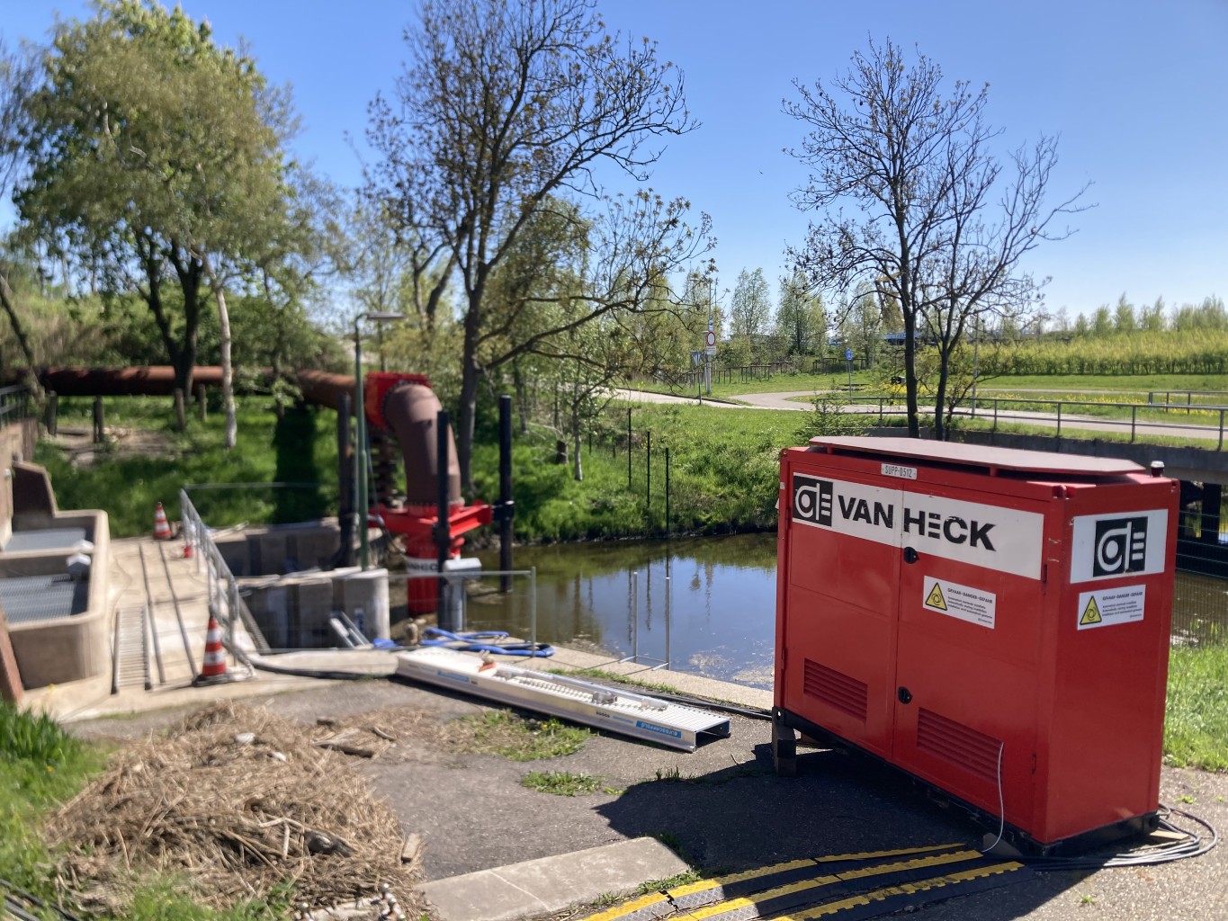 Tijdelijke Pompinstallatie bij gemaal Rode container met opschrift "Van Heck" met op de achtergrond een kanaal met daarin een rode pomp, in een groene omgeving met bomen