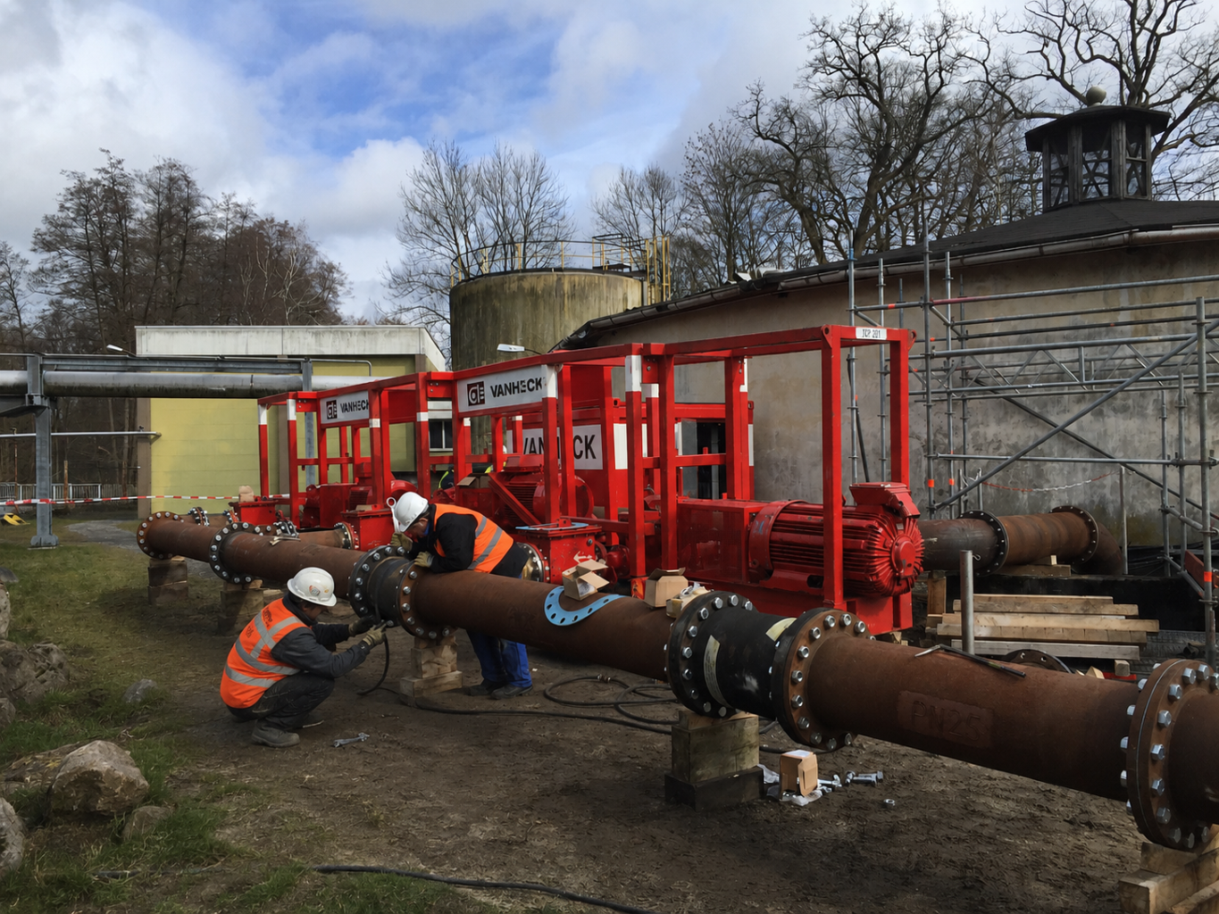 Installatie van een brandbluspompset twee monteurs werken aan leidingwerk op een industrieel terrein met drie rode waterpompen op de achtergrond