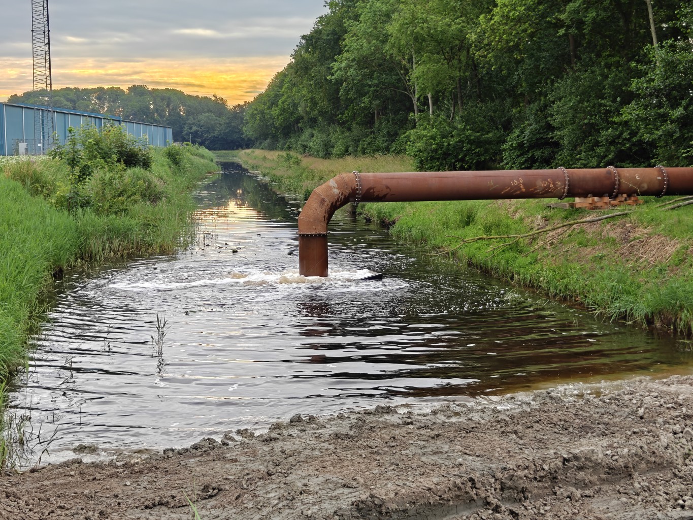 Stalen leiding in een sloot waar water uit komt in een groene omgeving met zonsondergang op de achtergrond