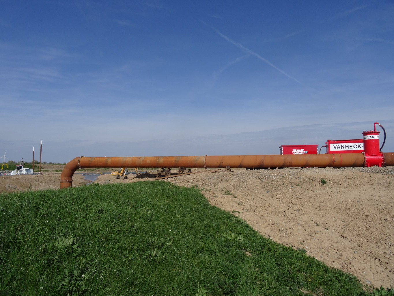 Stalen leiding met rode machines in een gras en zandlandschap onder een blauwe lucht