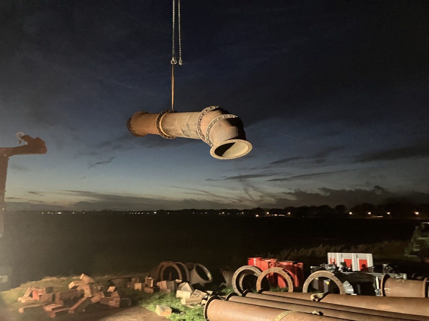Een stuk leidingwerk hangt in het donker in een kraan, op de grond liggen verschillende stukken leidingwerk.