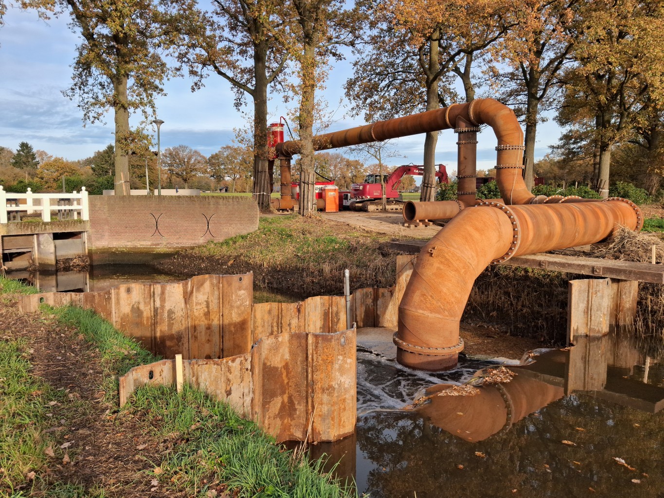 Hevel instroom Instroomzijde van een hevelinstallatie met twee damwanden in een watergang met bomen in herfstkleuren en een graafmachine op de achtergrond