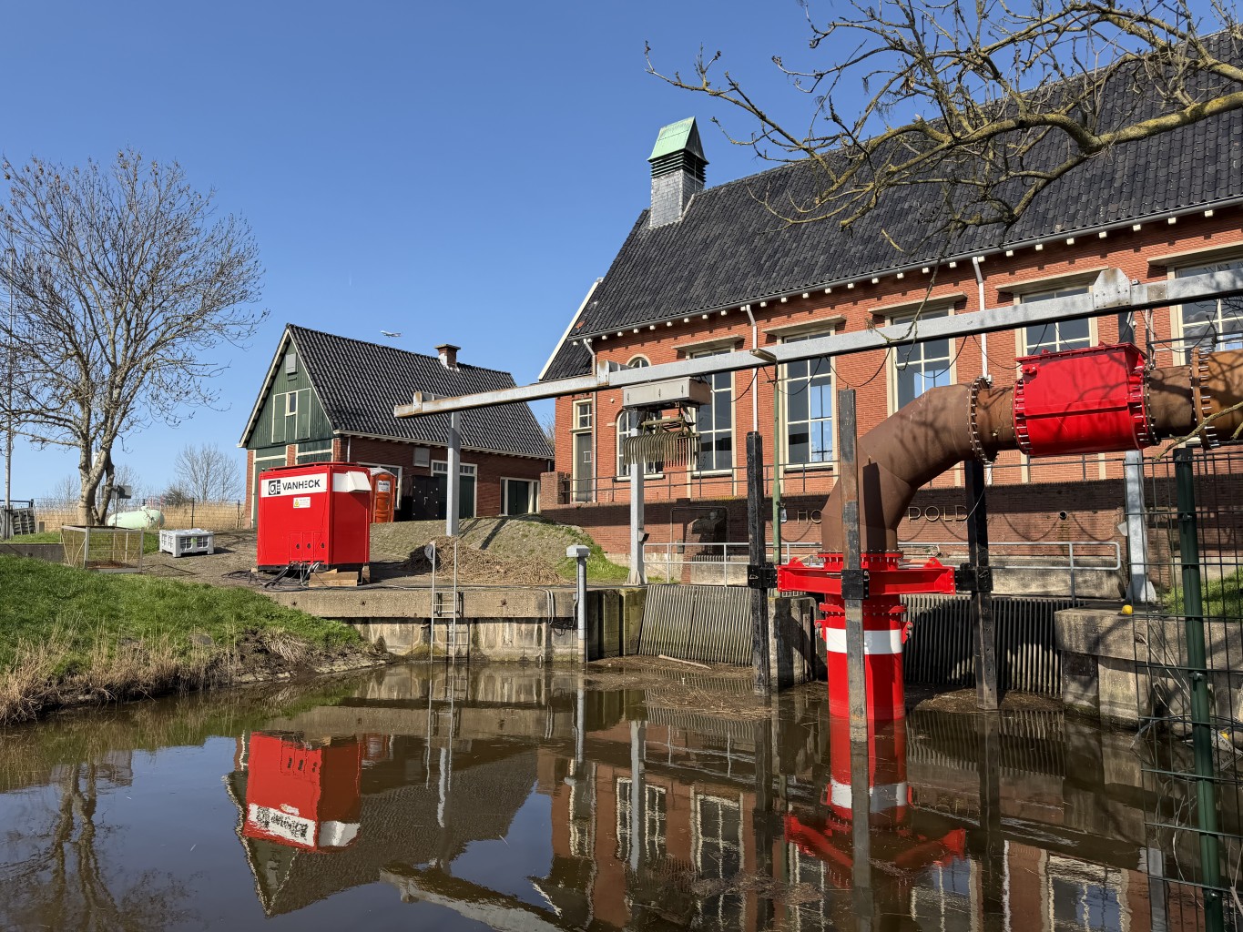 Tijdelijke pomp vp1200 bij een gemaal Rode pomp met leidingwerk in het water voor een gebouw met ramen en een huisje ernaast. Er staat links een rode container met het opschrift "van Heck"
