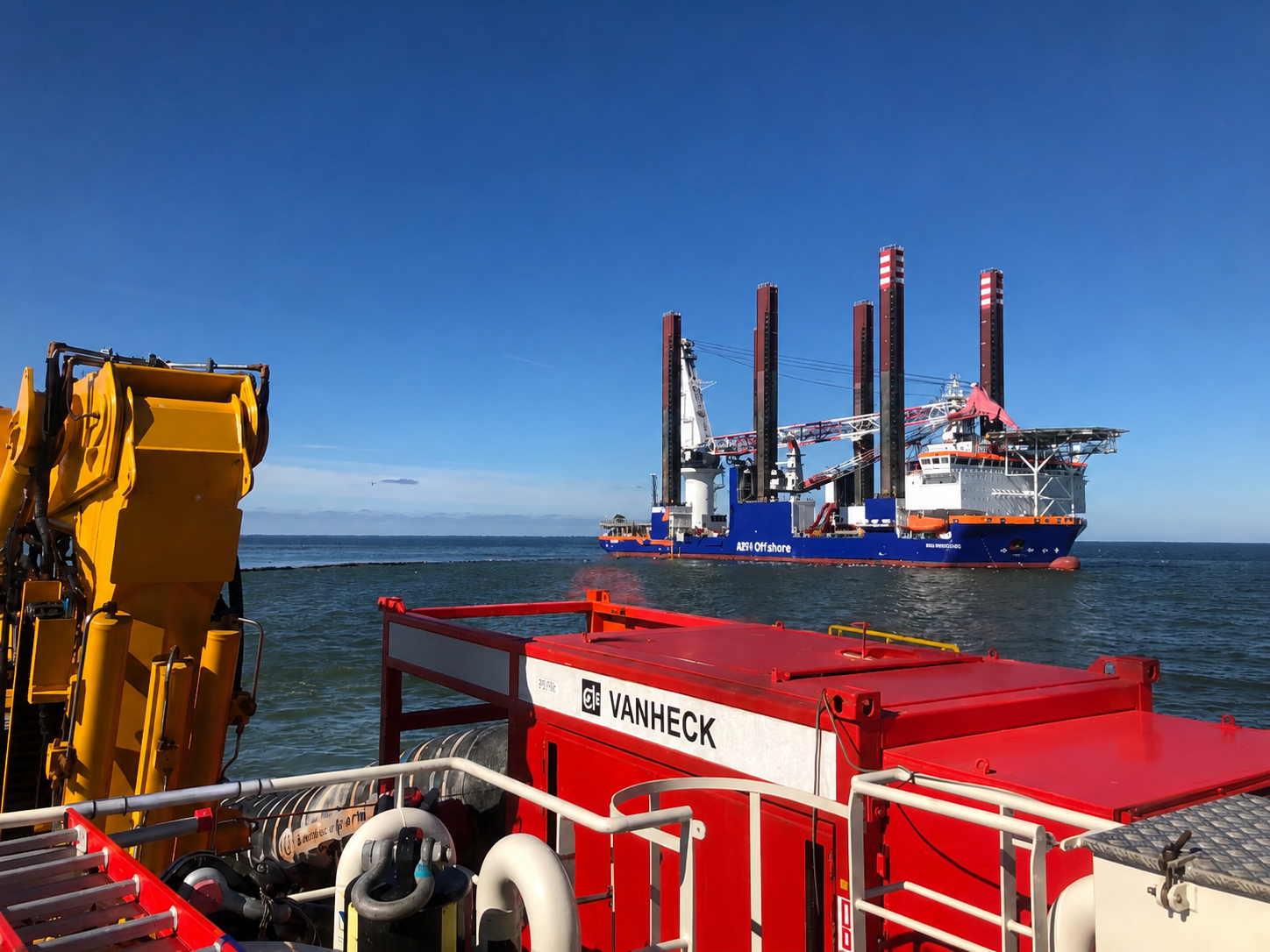 Hogedrukpomp op multicat Rode pomp op een multicat op zee, op de achtergrond is een blauw met oranje jack-up schip te zien