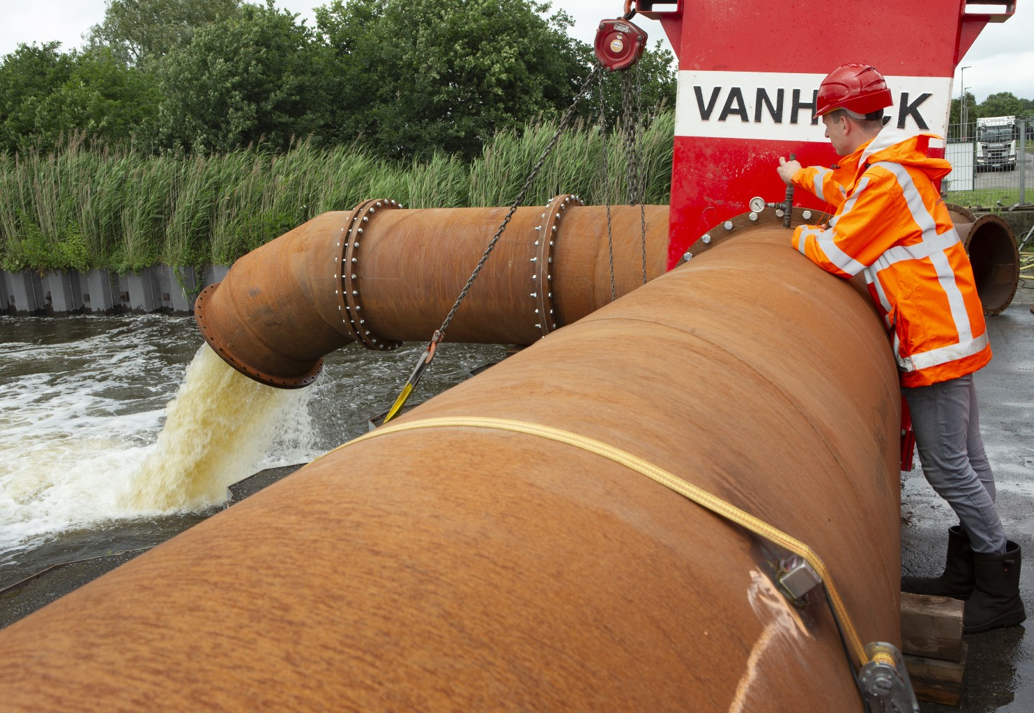 Testdraaien VP1200 Een monteur in signaalkleding leunt tegen een roestige stalen leiding voor een rode aflsuiter met de tekst "Van Heck" waar water uit stroomt in een kanaal. De achtergrond zijn groene bosjes